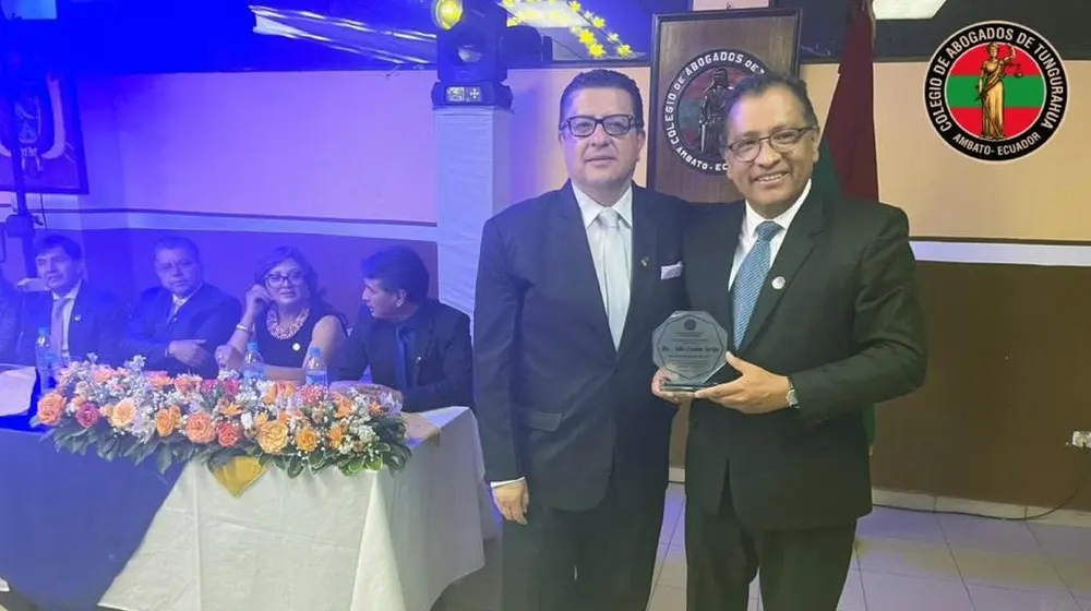 Entrega de un reconocimiento entre dos abogados frente a la mesa directiva decorada con flores