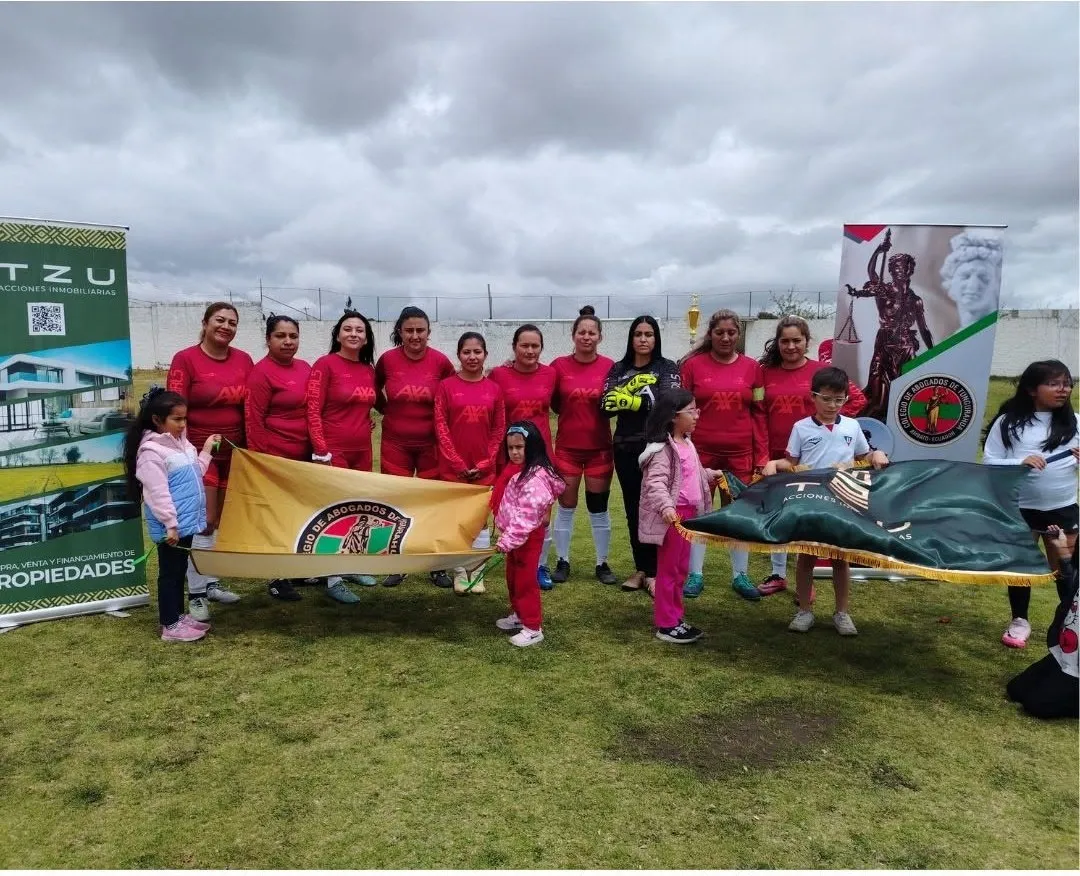 Equipo femenino de fútbol con camisetas rojas posa con la bandera del Colegio en la cancha