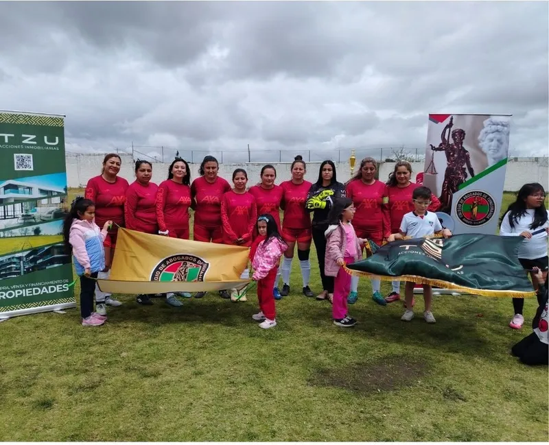 Equipo femenino de fútbol con camisetas rojas posa con la bandera del Colegio en la cancha