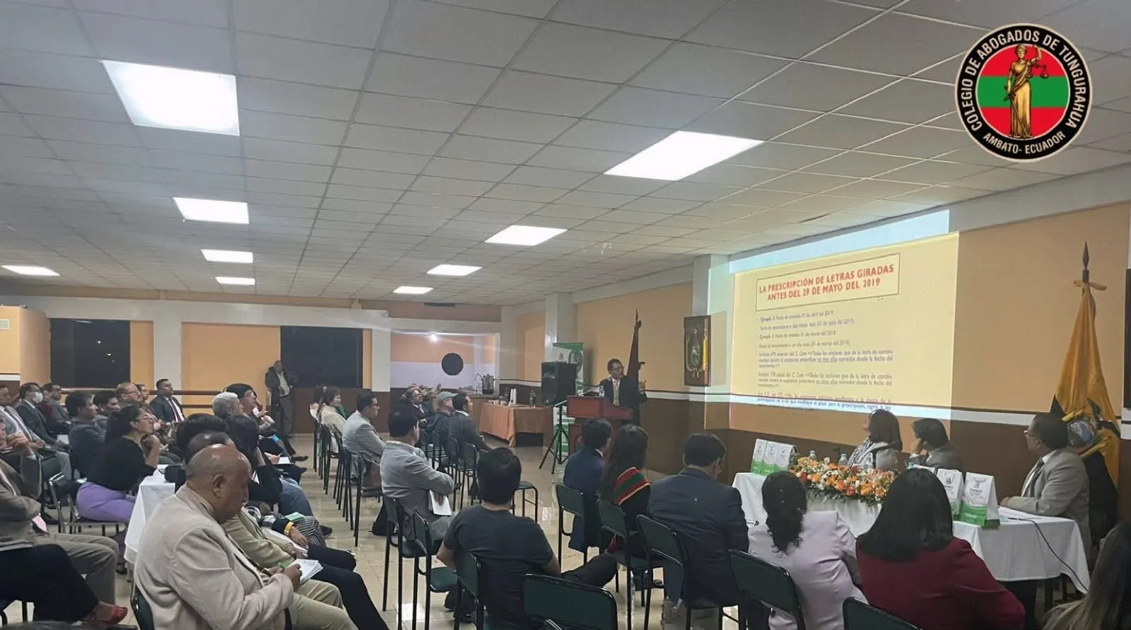 Asistentes sentados en el salón del Colegio durante un evento académico, con la mesa directiva y una presentación al fondo