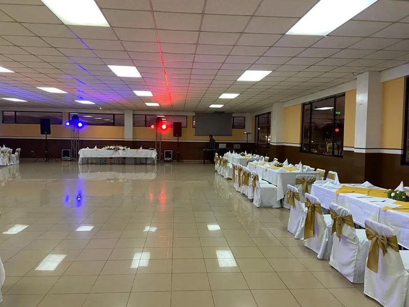 Salón de eventos amplio con mesas cubiertas de manteles blancos, sillas con lazos dorados, iluminación de techo y equipo de sonido